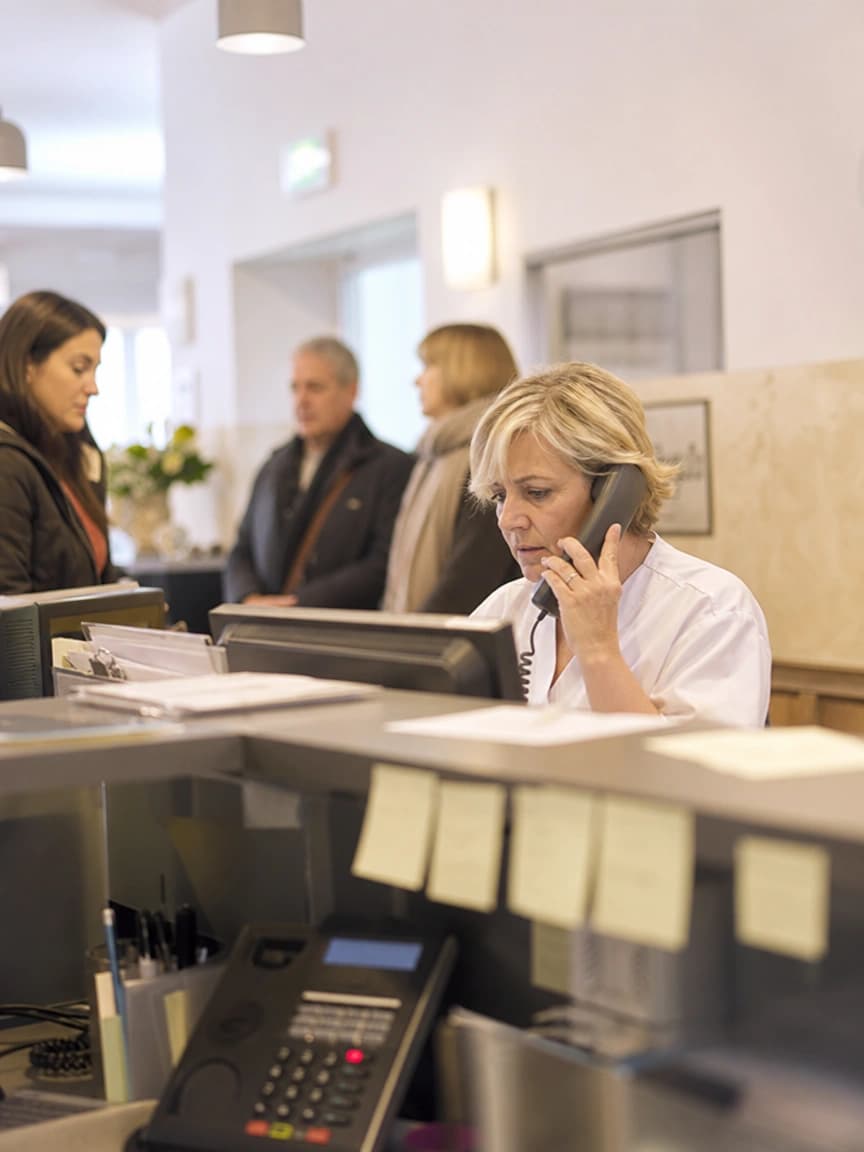 Segretaria di uno studio medico al telefono con un secondo apparecchio che squilla, mentre i pazienti aspettano
